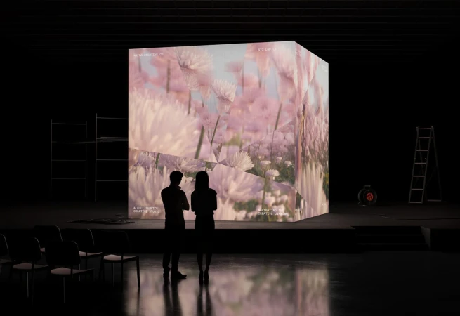 Two silhouetted figures stand before a large cube displaying a floral pattern in a darkened exhibition space with chairs and stage lighting.