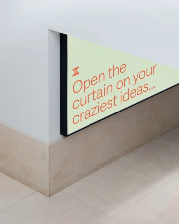 An interior wall featuring an illuminated, triangular sign with the text 'Open the curtain on your craziest ideas' in orange lettering.