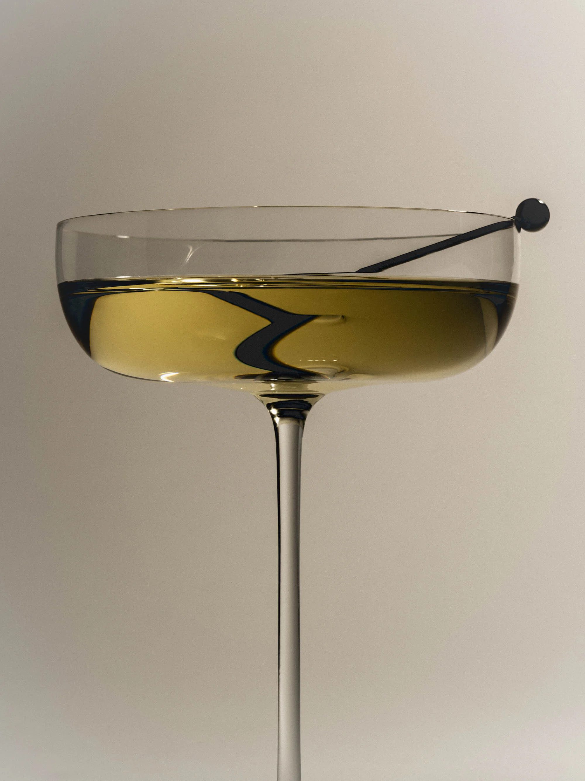 Elegant coupe glass filled with light-yellow liquid and black bar spoon against a cream-colored backdrop, creating a sophisticated composition.