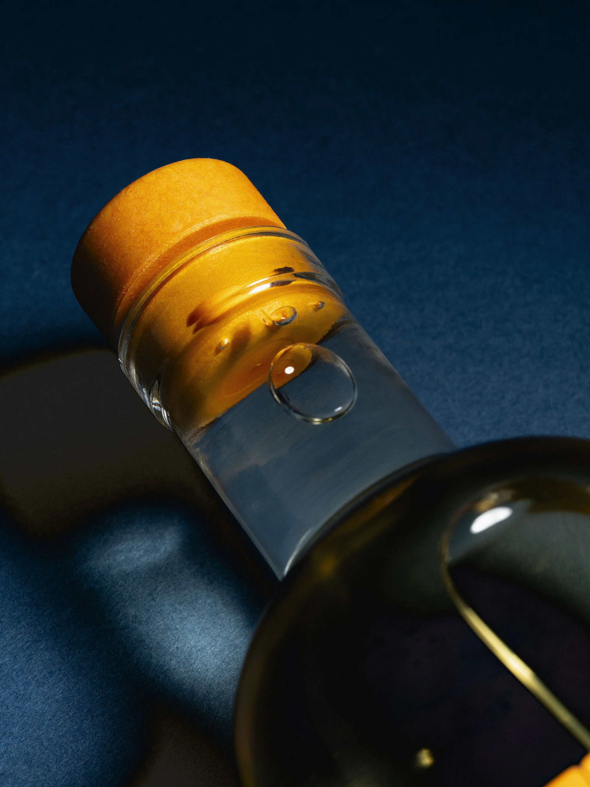 Close-up of a tilted clear glass bottle with liquid, sealed with an orange stopper, showcasing water droplets against a blue background.