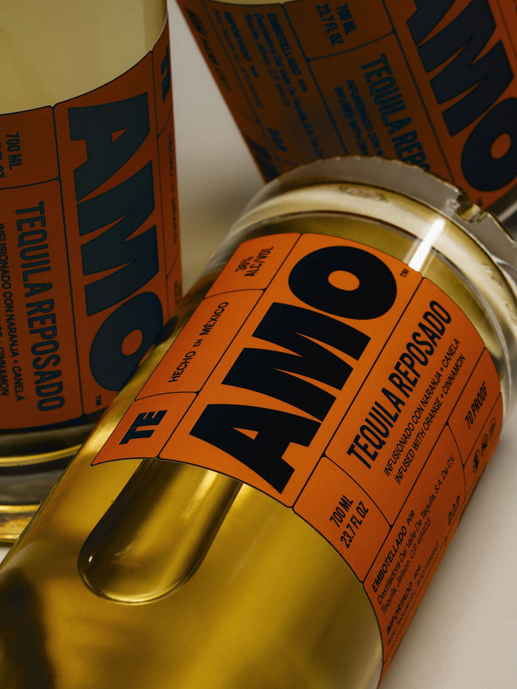 Close-up of three bottles of Tequila Reposado, featuring orange labels with bold black 'AMO' lettering. The bottles are slightly angled.