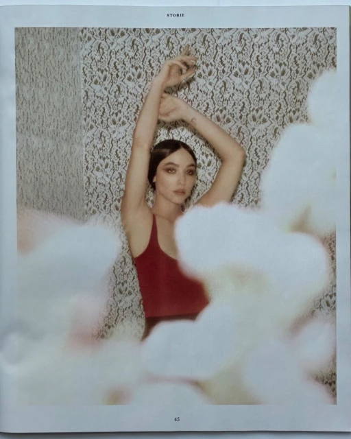 Young woman in red top poses with raised arms against a floral wallpaper, partially obscured by white flowers in the foreground.