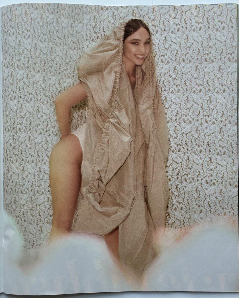 A smiling woman in an avant-garde hooded beige garment poses against a floral wallpaper backdrop, creating a striking fashion statement.