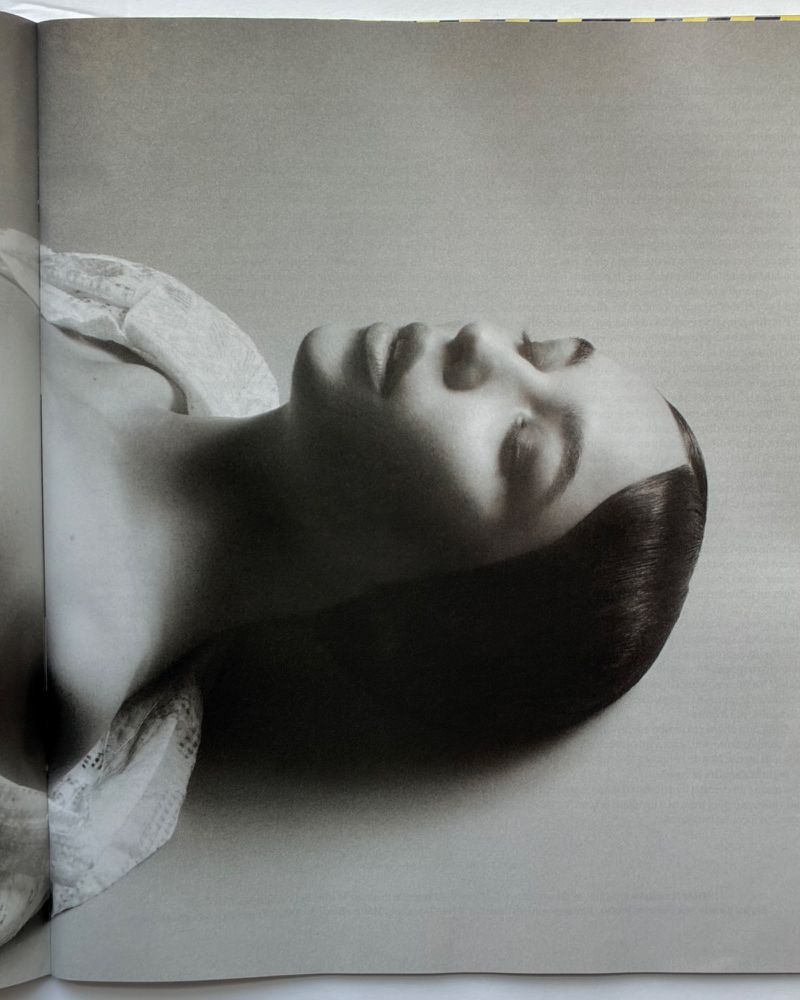 Monochrome portrait of a woman with closed eyes, lying down and resting her head on a surface, with a soft white fabric around her neck.