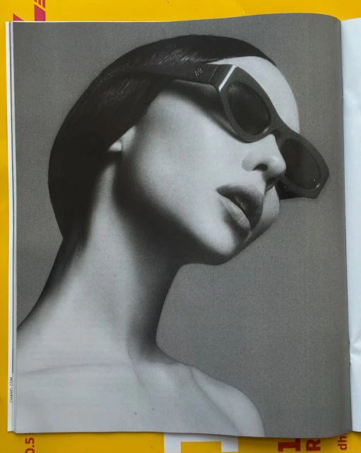 Black and white close-up of a woman with dark, slicked-back hair wearing geometric sunglasses, against a grey studio background.