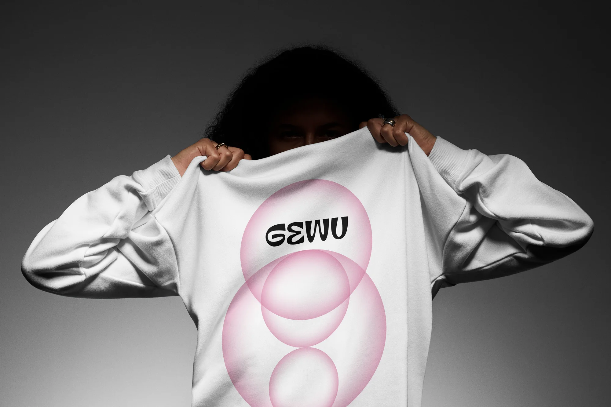 Person holding a white sweatshirt with overlapping pink circles and black text in a studio with gray gradient background.