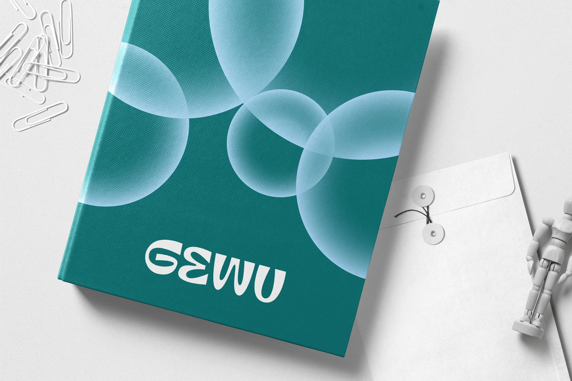 Close-up of a teal notebook with an abstract design, next to an envelope, paper clips, and an artist's figure on a white surface.