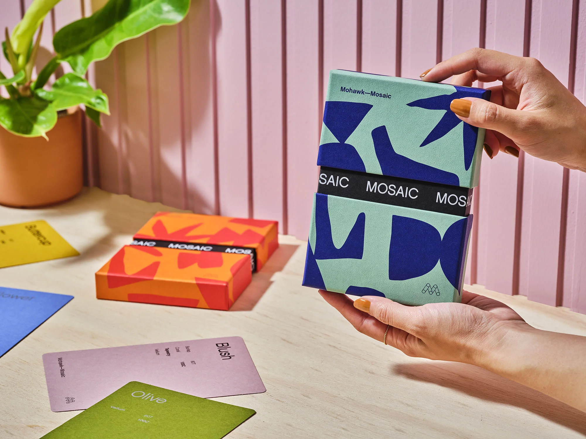 Hands hold a teal and navy Mohawk Mosaic box with orange boxes and colorful cards on a light wood table with a pink and white wall.