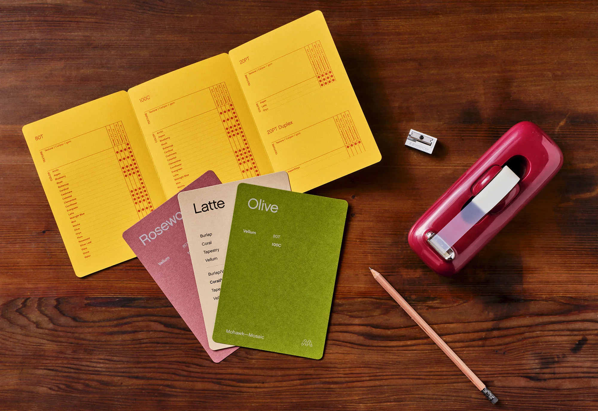 Open yellow notebook with color swatch cards, pencil, metal sharpener, and red eraser on dark wooden surface in flat lay style.