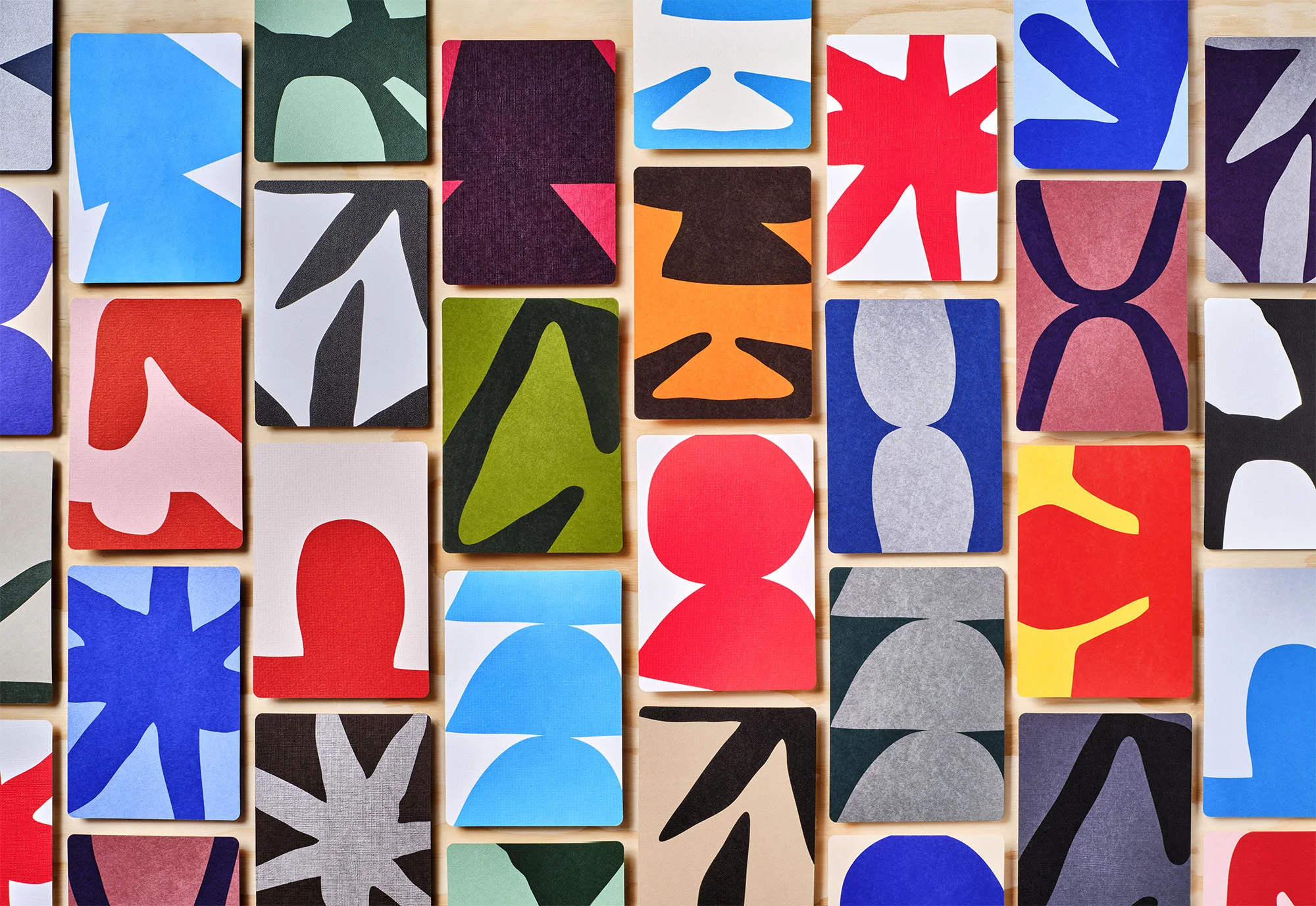 Colorful collection of graphic cards with geometric patterns and shapes in red, blue, green, black, and white on a light wood background.