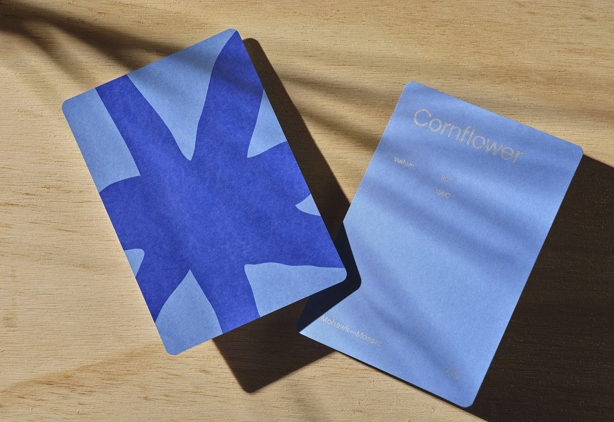 Two cards, one with a blue abstract floral design, and the other with the word Cornflower on a light blue surface, sit on a wood surface.