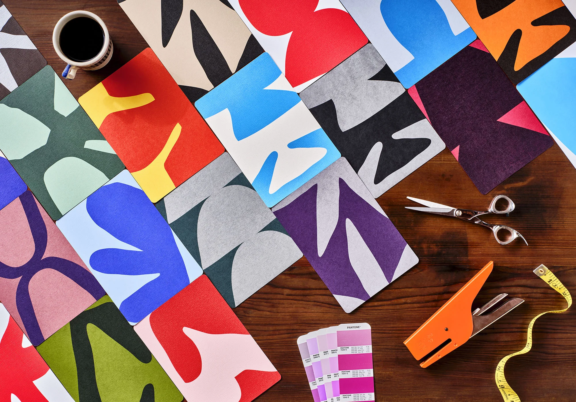 Overhead shot of colorful paper cutouts arranged on a wooden surface, complemented by scissors, a stapler, measuring tape, and a cup of coffee.