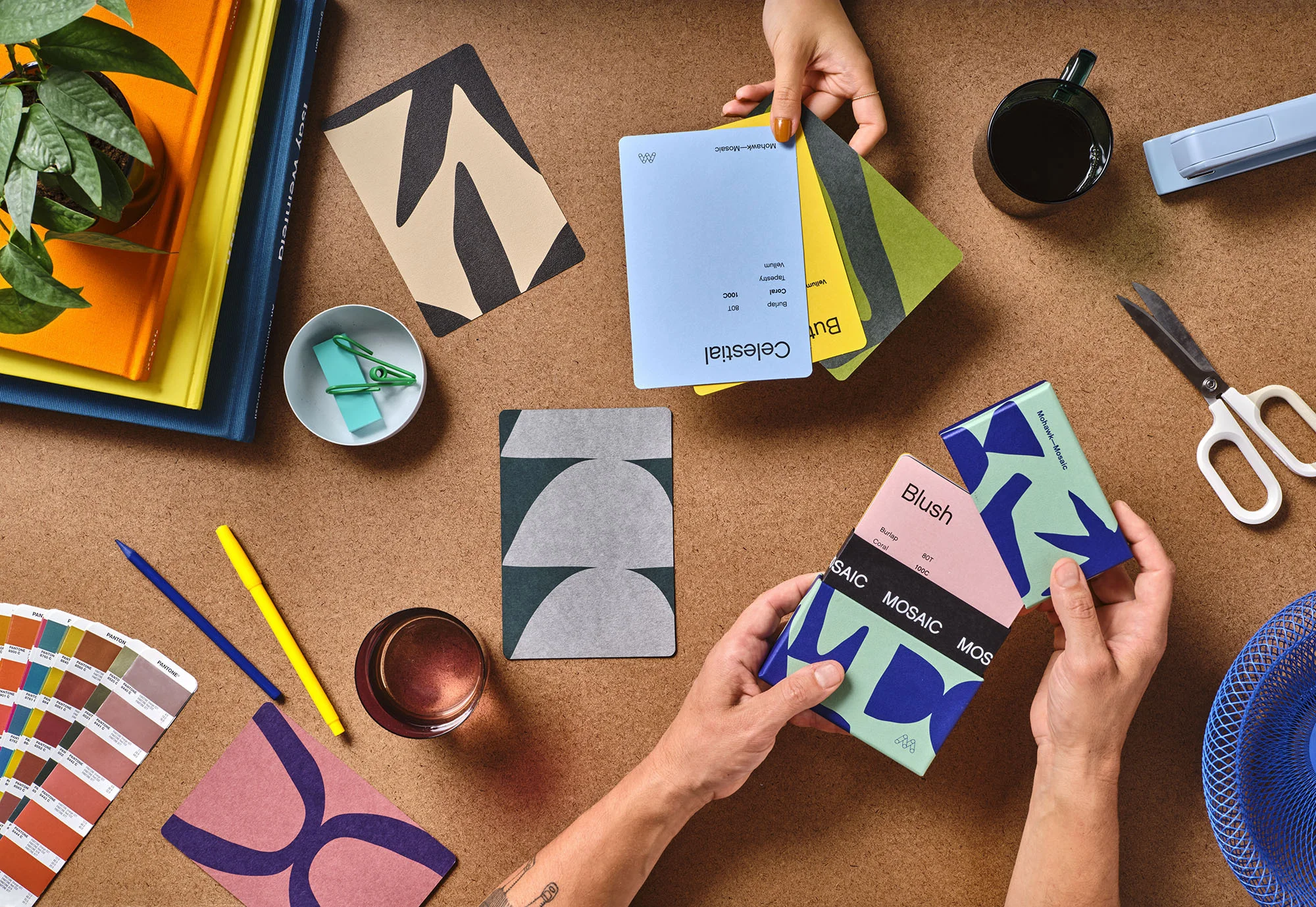 Overhead shot of hands arranging color palette cards, books, pencils, scissors, and a drink on a brown textured surface.