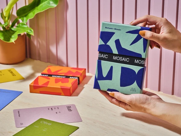 Hands holding a teal and blue patterned paper box for Mohawk Mosaic on a wooden surface with a pink paneled wall.