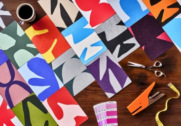 Flat lay of colorful abstract paper art cards and crafting tools on a dark wooden table with a coffee cup.