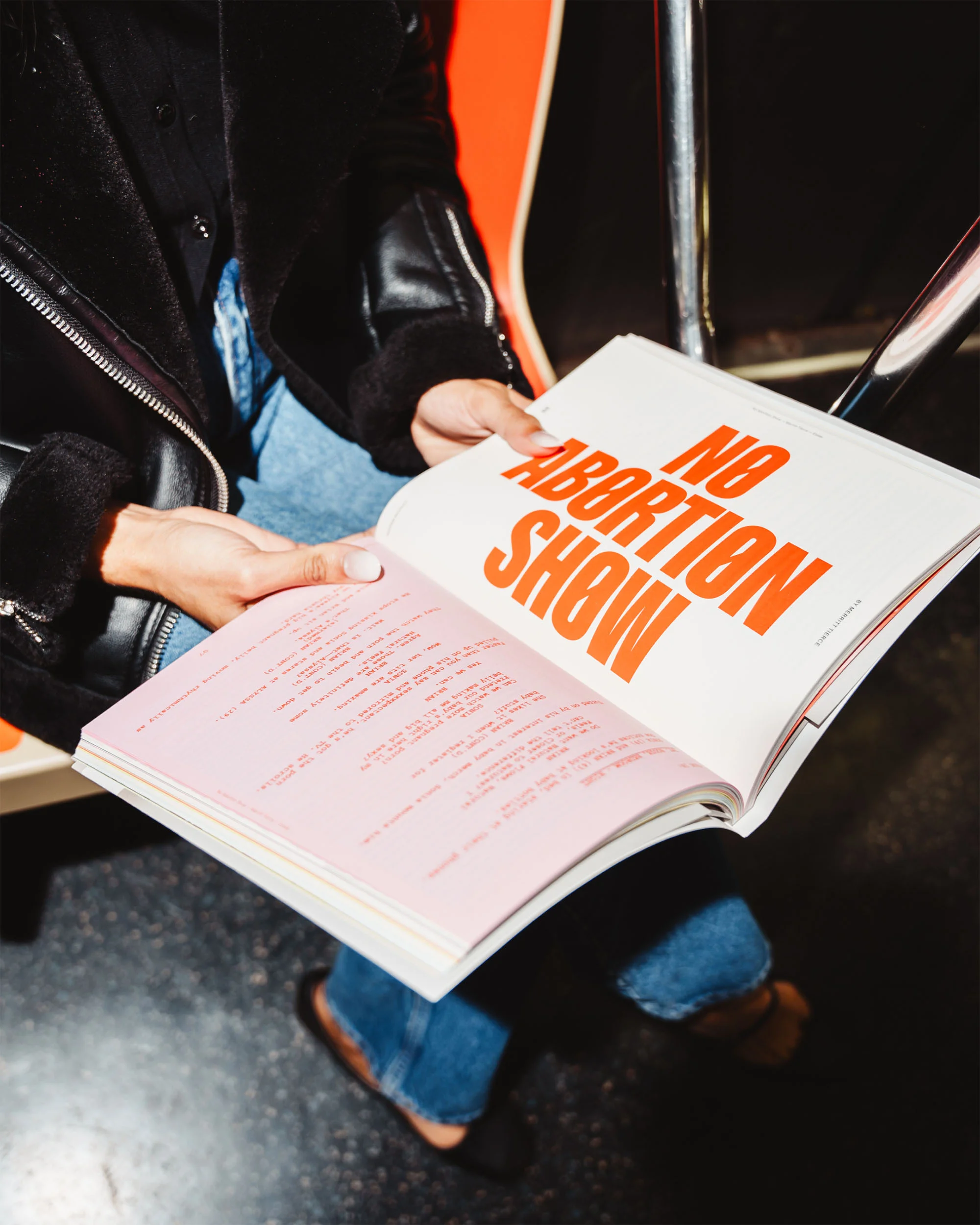 Person reading 'No Abortion Show' on public transport, wearing a jacket, jeans, and flats. The book's cover is white with orange text.