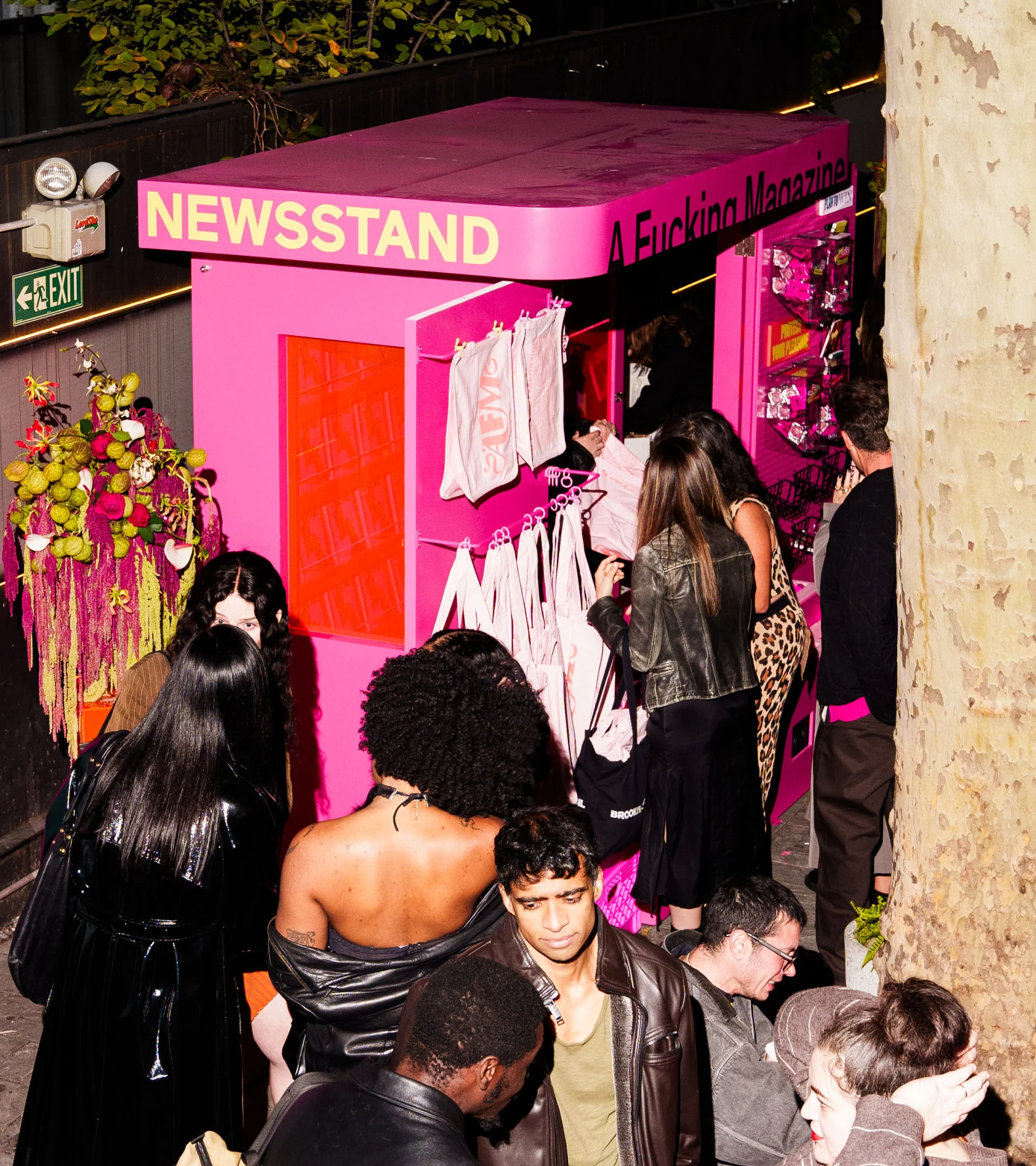 Pink newsstand, surrounded by a diverse crowd of people, displays magazines and tote bags in an outdoor urban setting, captured at eye-level.