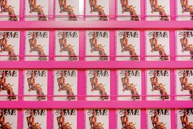 Multiple identical copies of a magazine displayed on bright pink display shelves with bold typographic covers