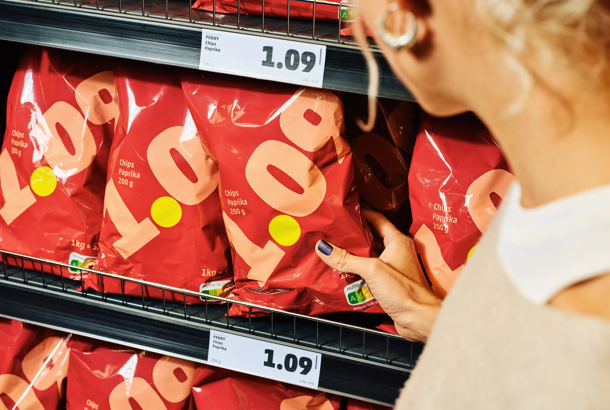 Woman selects a bag of paprika chips from a shelf in a grocery store. Red packaging with yellow accents on a dark metal shelf.