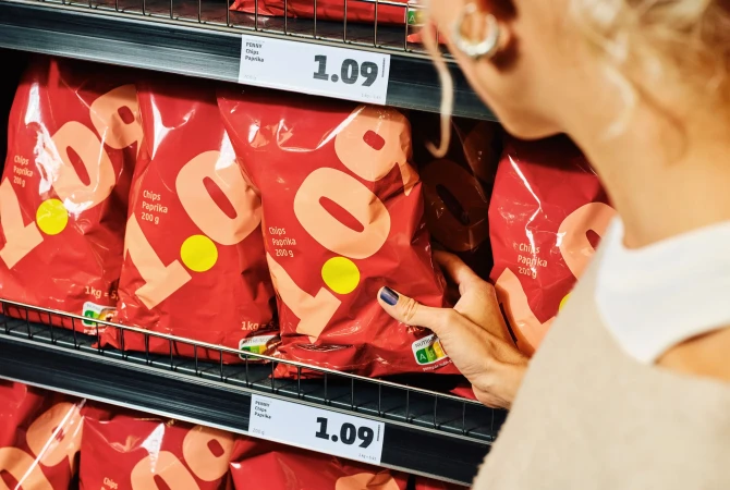 Woman selects a bag of paprika chips from a shelf in a grocery store. Red packaging with yellow accents on a dark metal shelf.