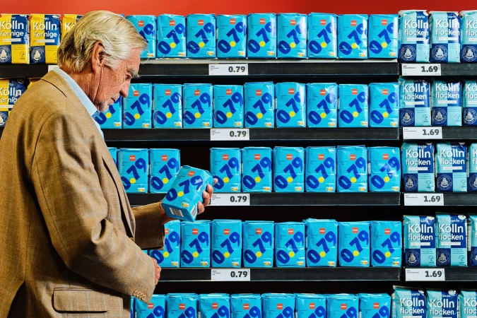 Elderly man in a tan jacket selecting a box of Kölln oat flakes from a shelf in a store, surrounded by numerous identical boxes.