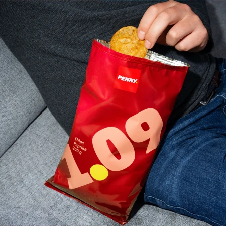 Person reaching into a red bag of Penny brand paprika chips while sitting on a grey couch in casual attire, promoting snack indulgence.