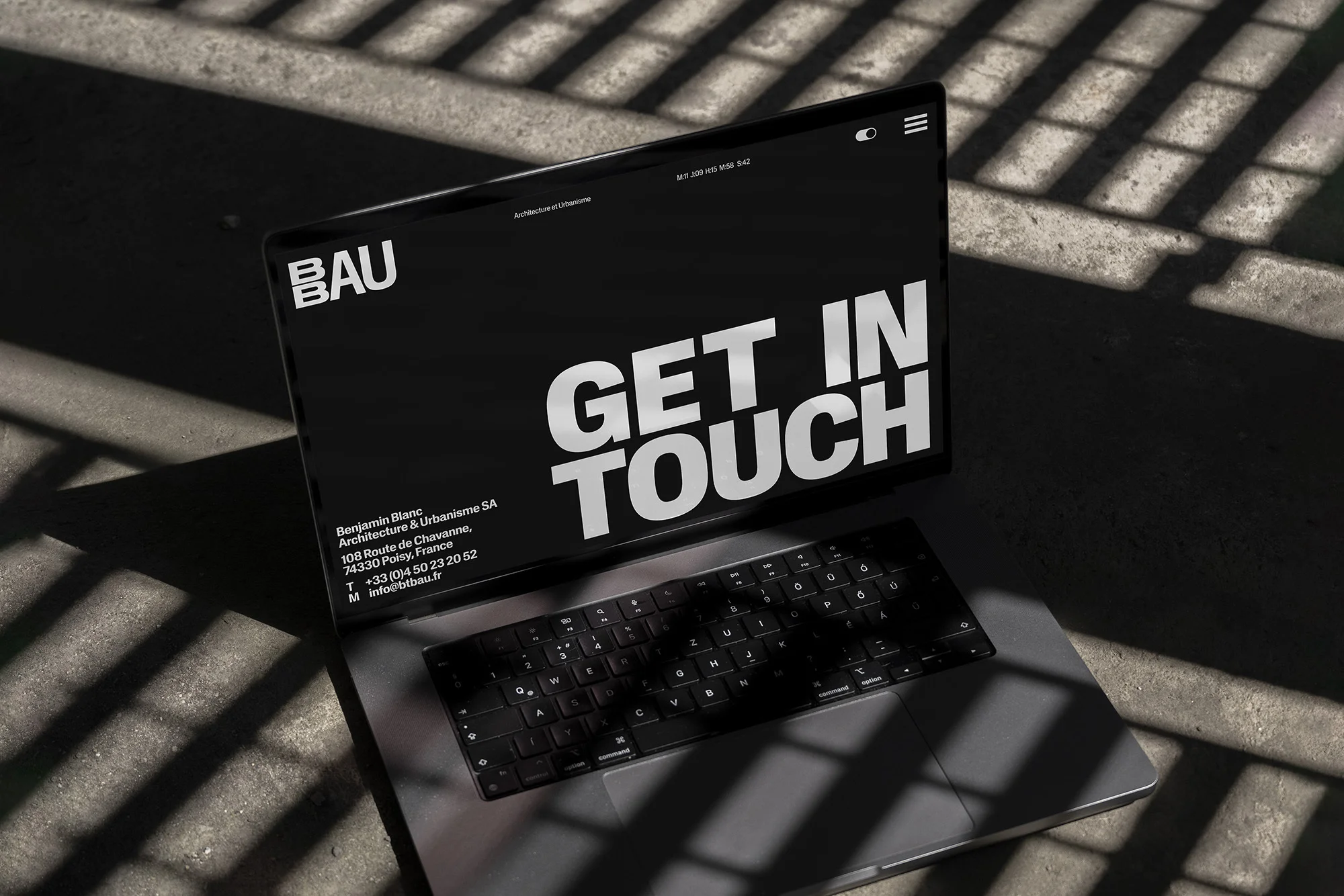 Laptop displays architecture website with bold 'GET IN TOUCH' text, set on a concrete surface with a play of light and shadow.