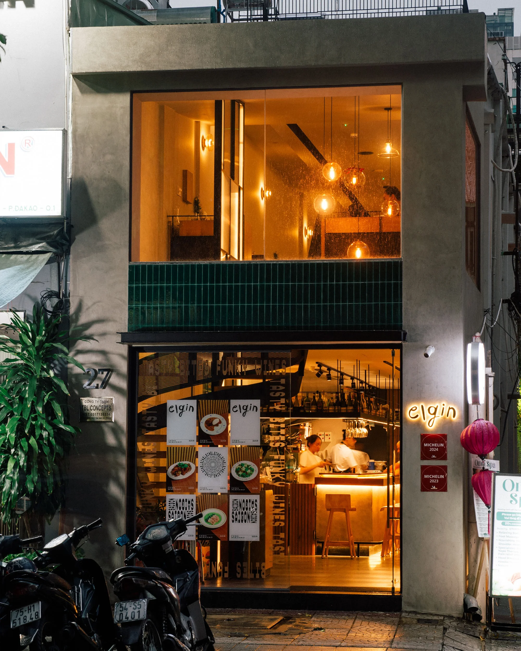 Exterior view of Elgin restaurant at night with visible interior featuring bar, seating, and staff, scooters parked outside.