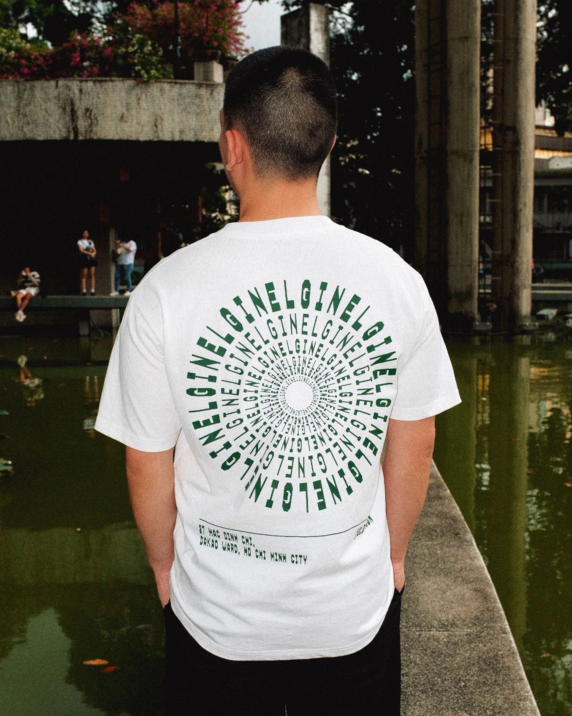 Man wearing a white t-shirt with a circular green graphic design, standing outdoors near a pond and greenery in an urban park setting.