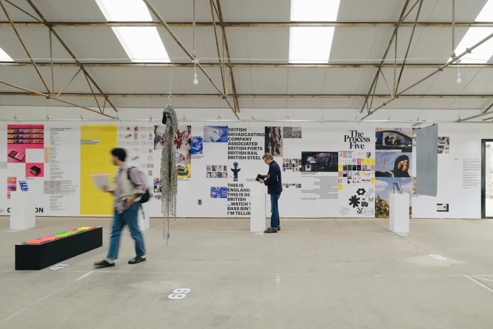 Two people view art on display in an airy warehouse-like exhibition space with posters, sculptures, and bright neon colors on display.
