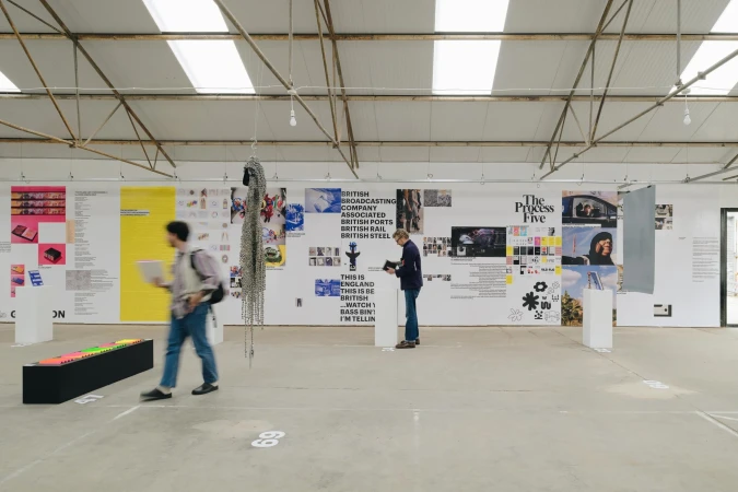 Two people view art on display in an airy warehouse-like exhibition space with posters, sculptures, and bright neon colors on display.