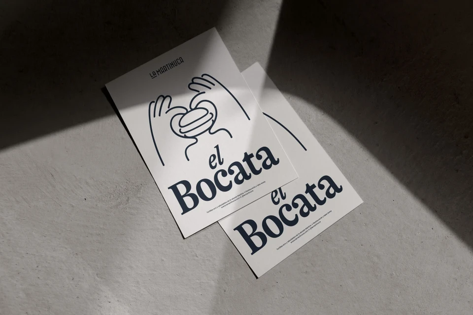Two identical flyers advertise 'el Bocata' with a cartoon sandwich graphic, lying on a textured gray surface in soft, diffused light.