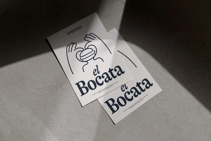 Two identical flyers advertise 'el Bocata' with a cartoon sandwich graphic, lying on a textured gray surface in soft, diffused light.