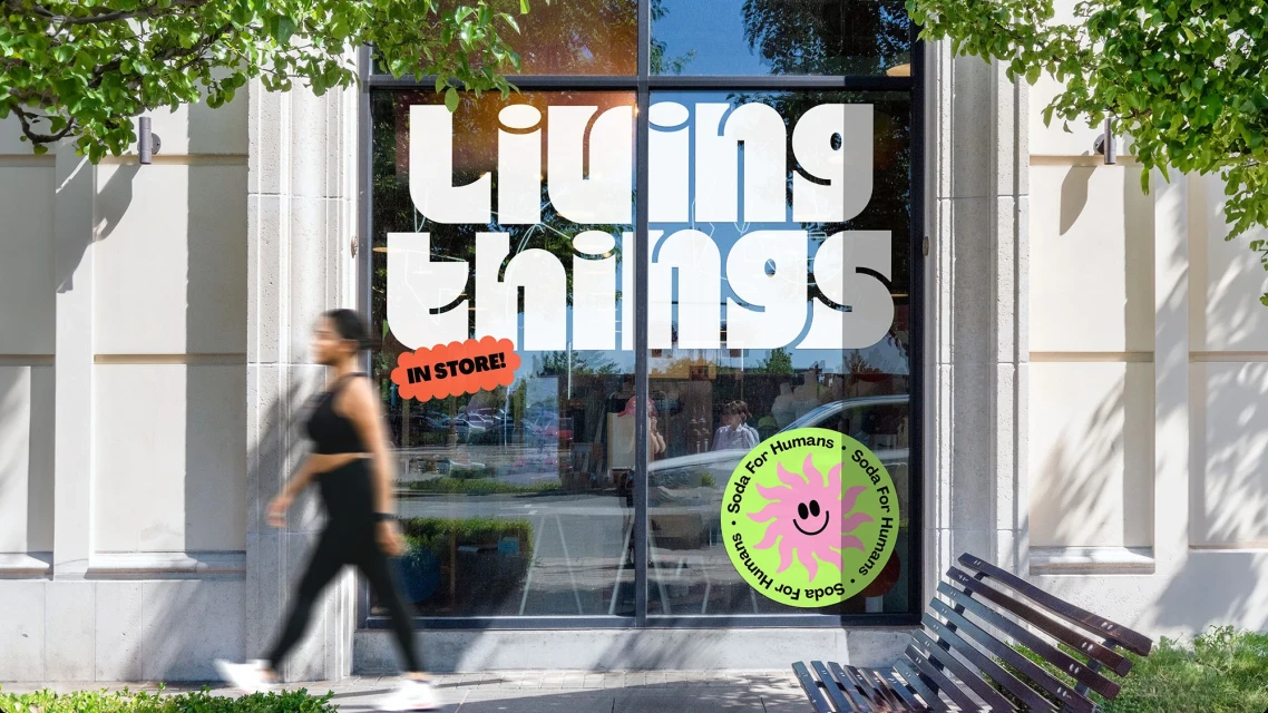 Woman walks by a storefront with 'Living Things IN STORE!' signage and a bright 'Soda for Humans' logo, framed by green tree branches.