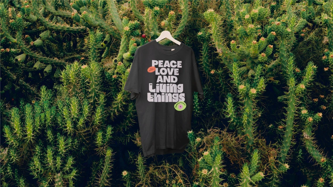 Black t-shirt on a hanger with 'Peace Love and Living Things' text, displayed against a vibrant green cactus background, outdoor setting.