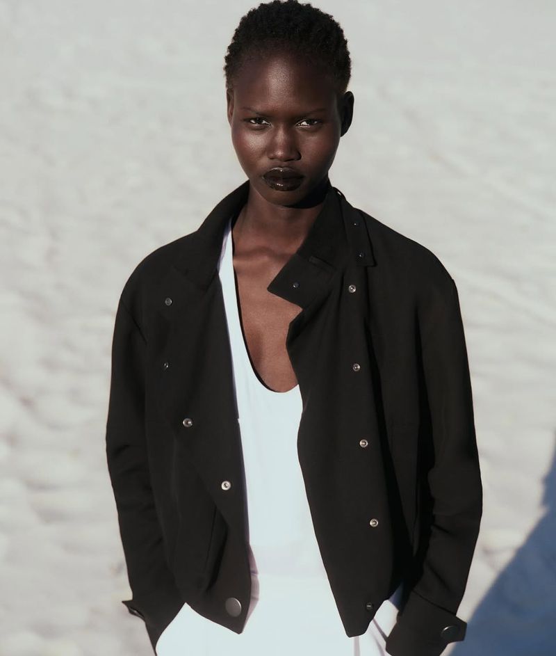 Portrait of a black female model with short hair wearing a black jacket over a white top, against a blurred white background.