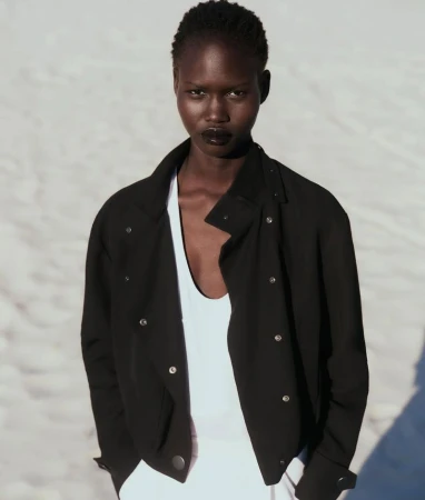 Portrait of a black female model with short hair wearing a black jacket over a white top, against a blurred white background.