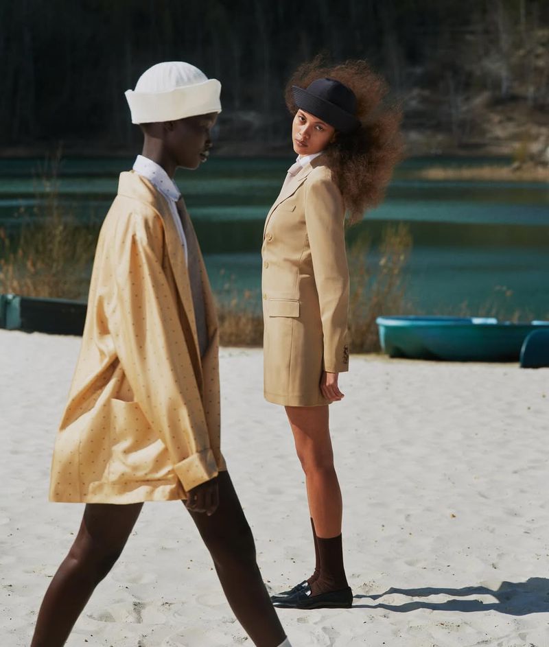 Two stylish models pose on a sandy beach near a lake. One wears a yellow jacket and hat while the other is in a tan suit.