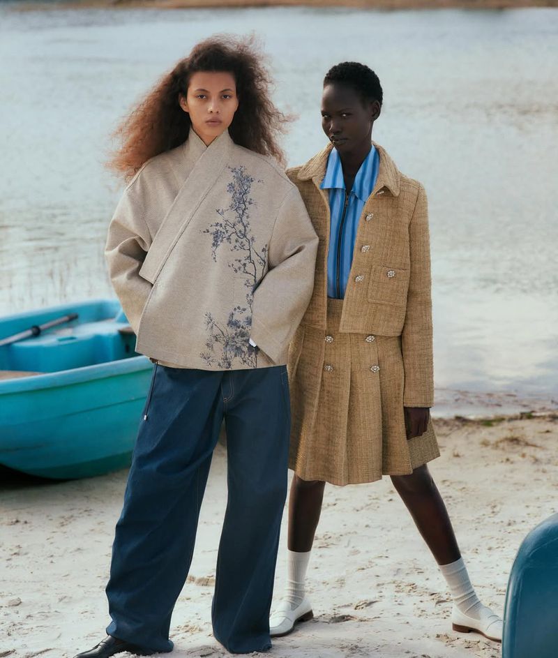 Two women model outdoor fashion. One wears a beige jacket with a floral design, dark pants. The other wears a tan skirt suit.