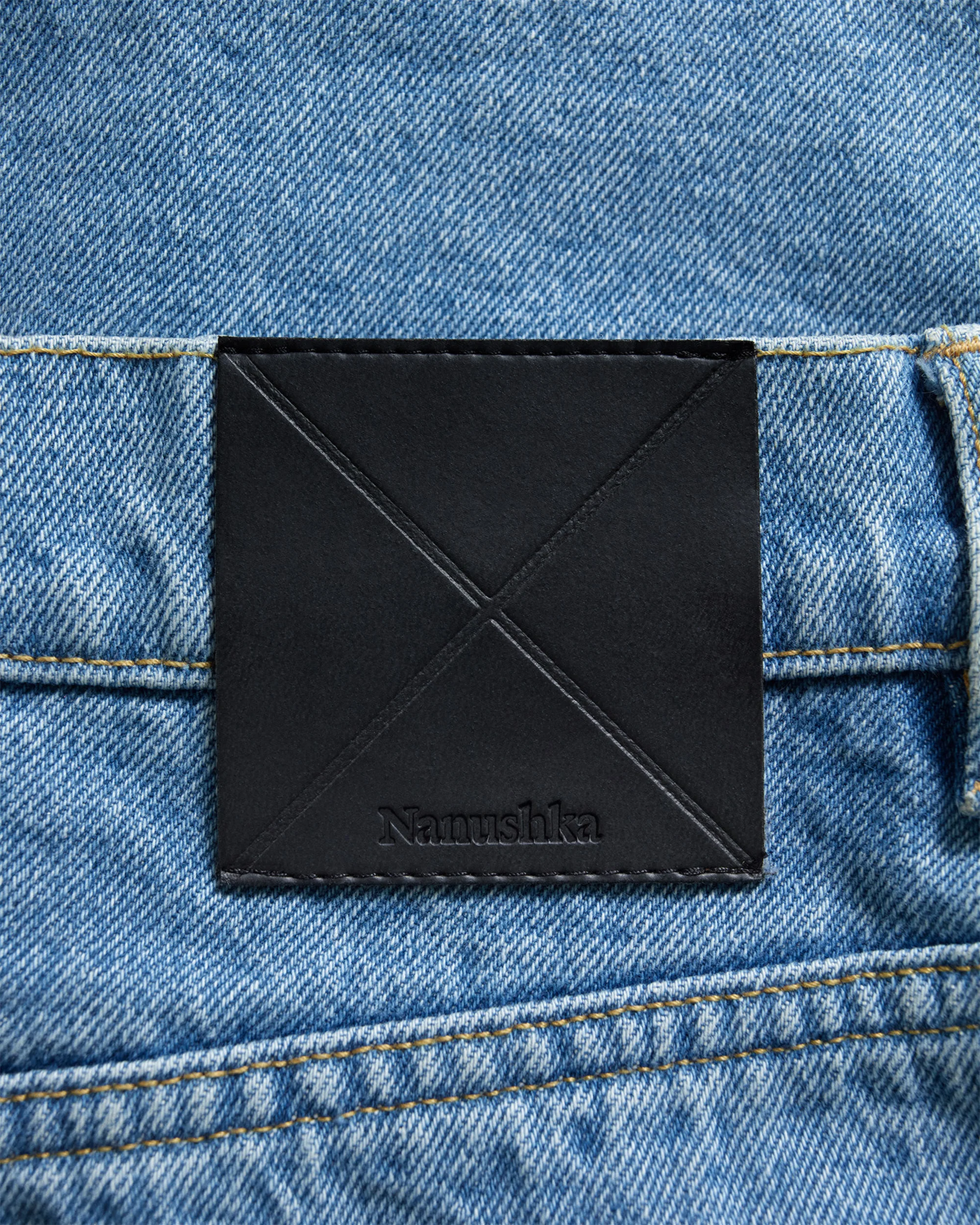 Close-up of light blue denim jeans with a black leather logo patch featuring stitching detail and the 'Nanushka' brand name.