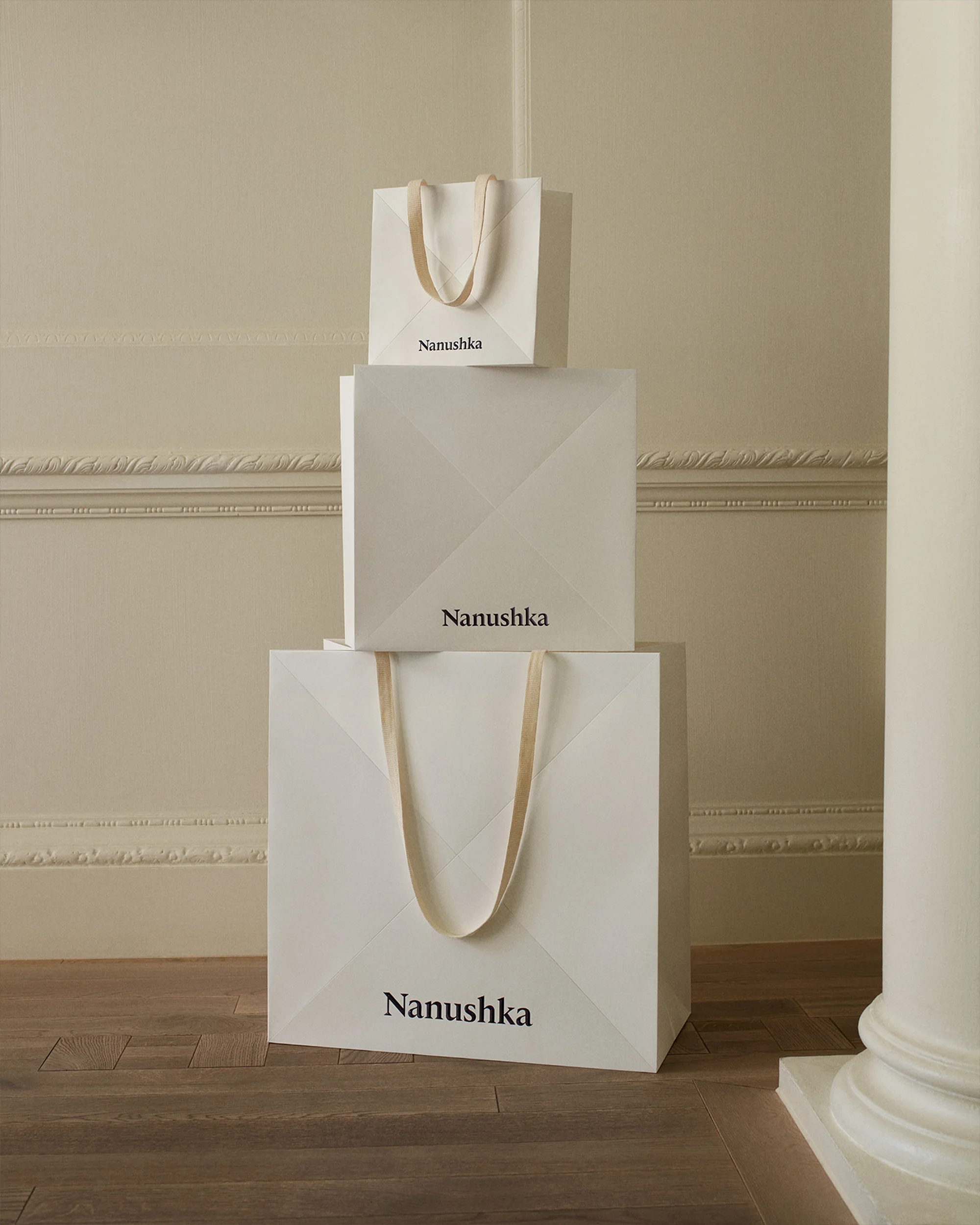 Three white Nanushka branded shopping bags stacked on top of each other on a wooden floor against a cream colored wall with molding.