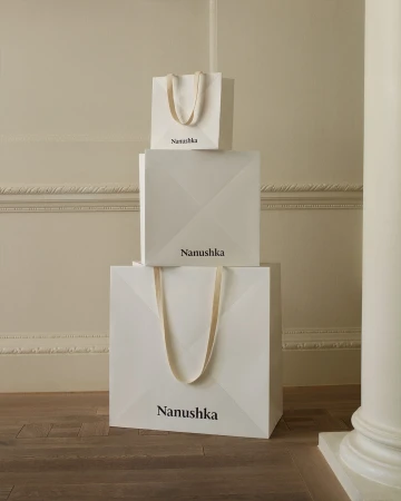 Three white Nanushka branded shopping bags stacked on top of each other on a wooden floor against a cream colored wall with molding.