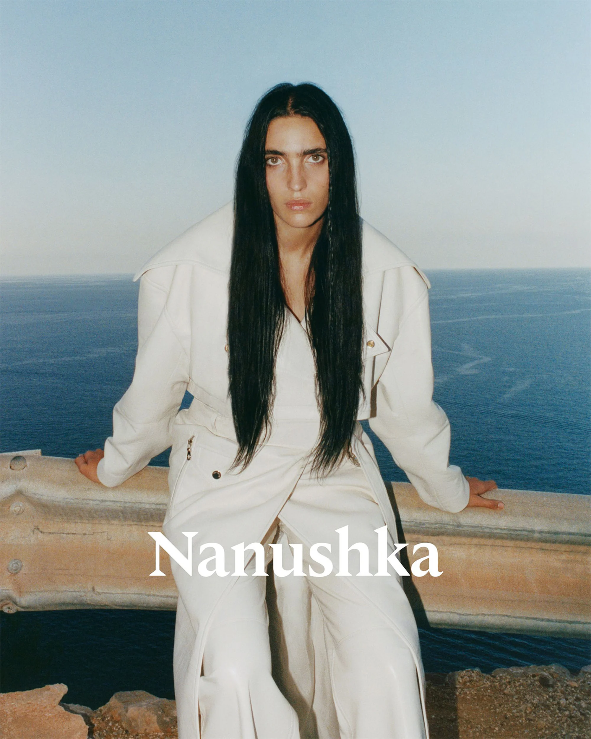 Fashion shot of woman in white jacket and pants standing by a ledge with ocean and sky background, Nanushka brand logo visible.
