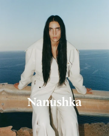 Fashion shot of woman in white jacket and pants standing by a ledge with ocean and sky background, Nanushka brand logo visible.