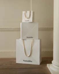 Three stacked white branded Nanushka shopping bags with ribbon handles in an interior room