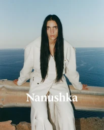 Woman with long dark hair wearing a white coat leaning against a roadside barrier overlooking the ocean.