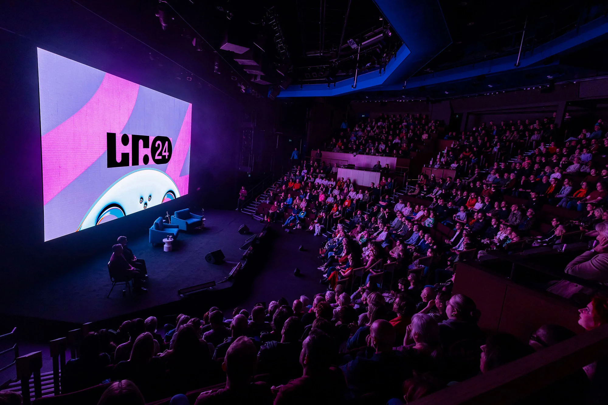 Audience watches a screen and presenters on stage in an indoor theater setting, bathed in blue and purple hues with stage lighting.