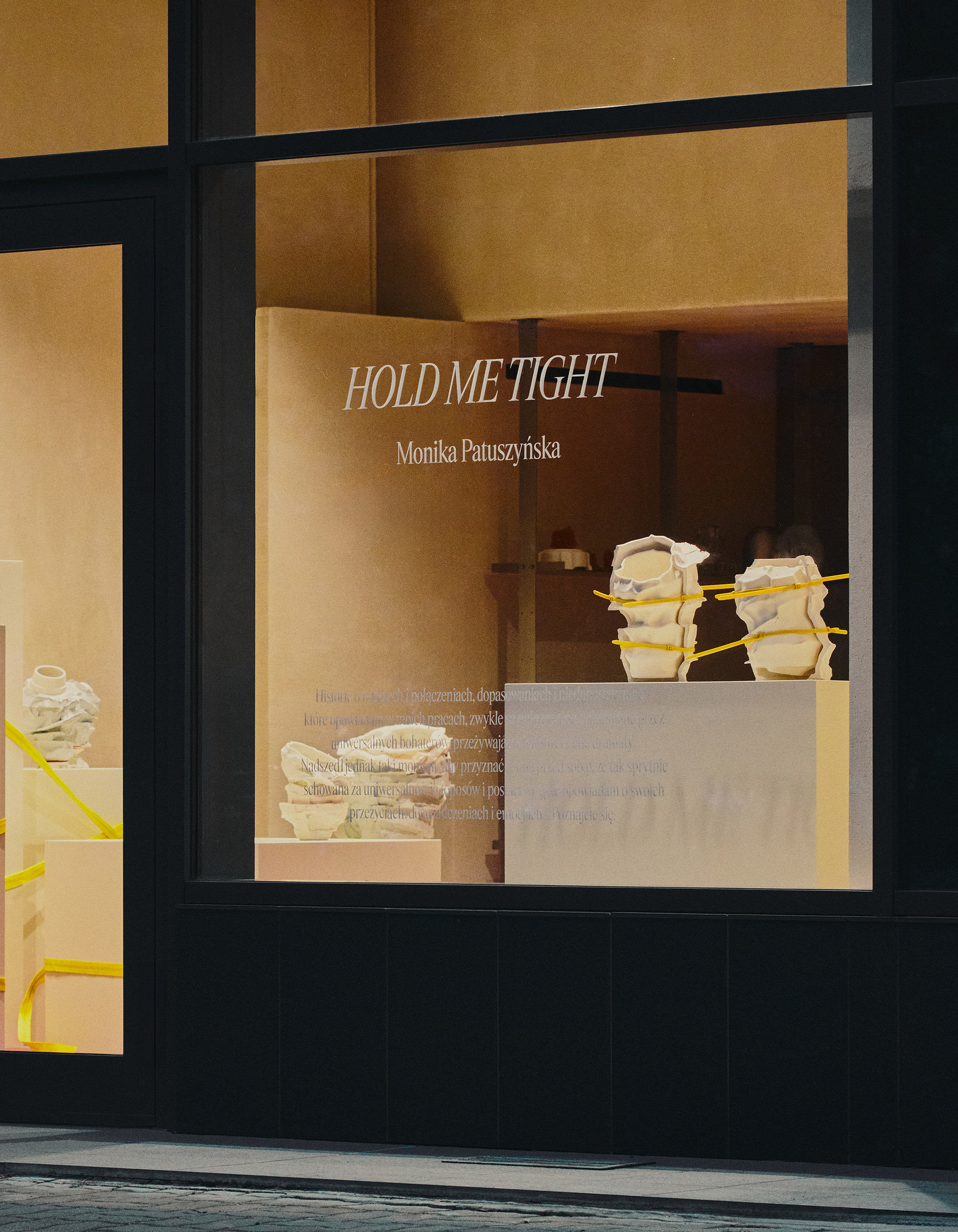 Storefront window displays ceramic sculptures by Monika Patuszyńska on pedestals, illuminated by warm lighting with 'Hold Me Tight' text.