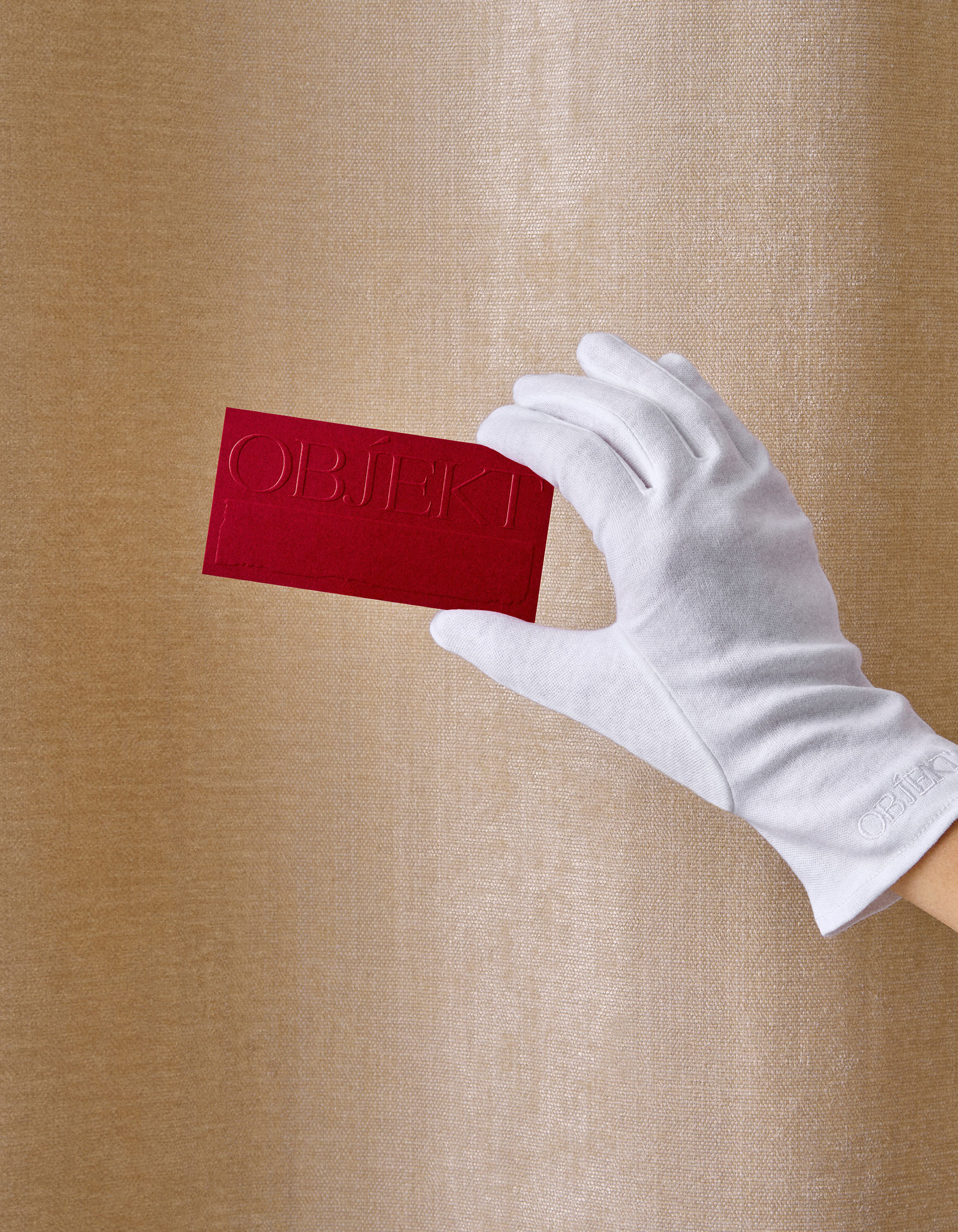 Hand in white glove holds a red card with the word OBJEKT against a textured gold background, creating a striking contrast.
