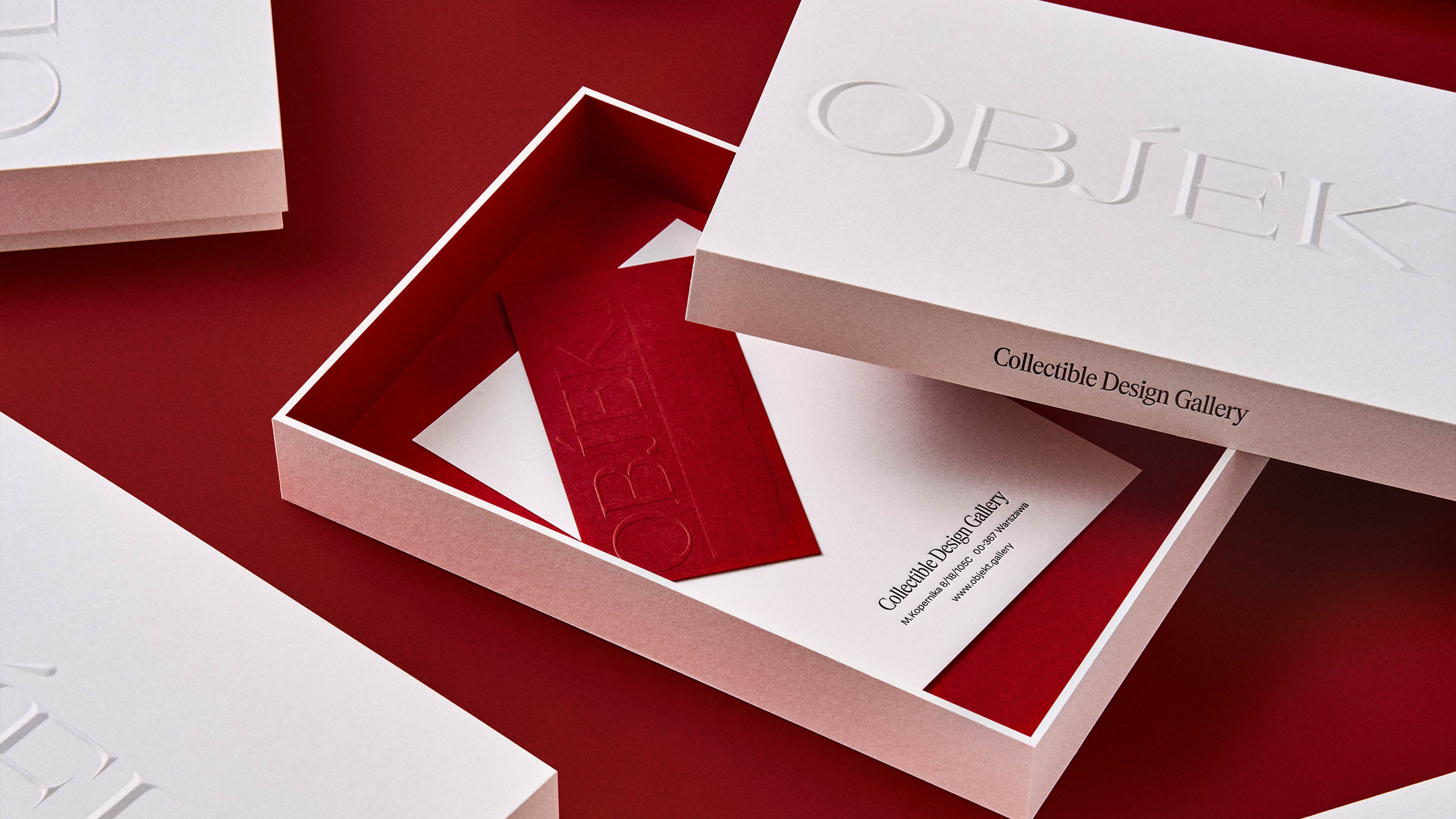 High-angle shot of a white, square box with an open lid, revealing a red interior and a card, against a red surface.